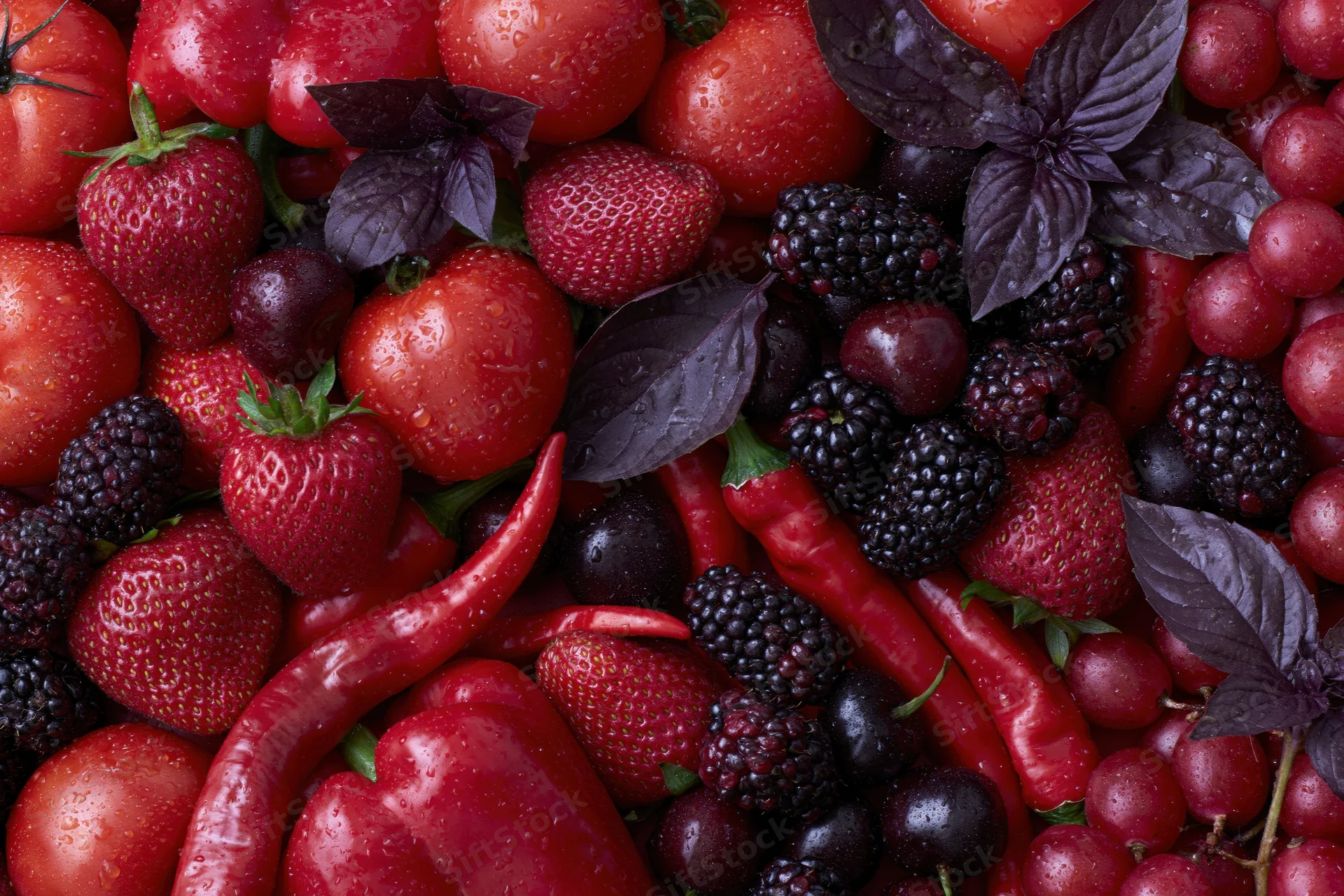 Overhead flat lay of mixed red and deep purple fruits and vegetables with basil leaves and water droplets