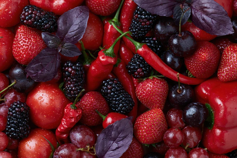 Overhead flat lay of mixed red and deep purple fruits and vegetables with basil leaves and water droplets