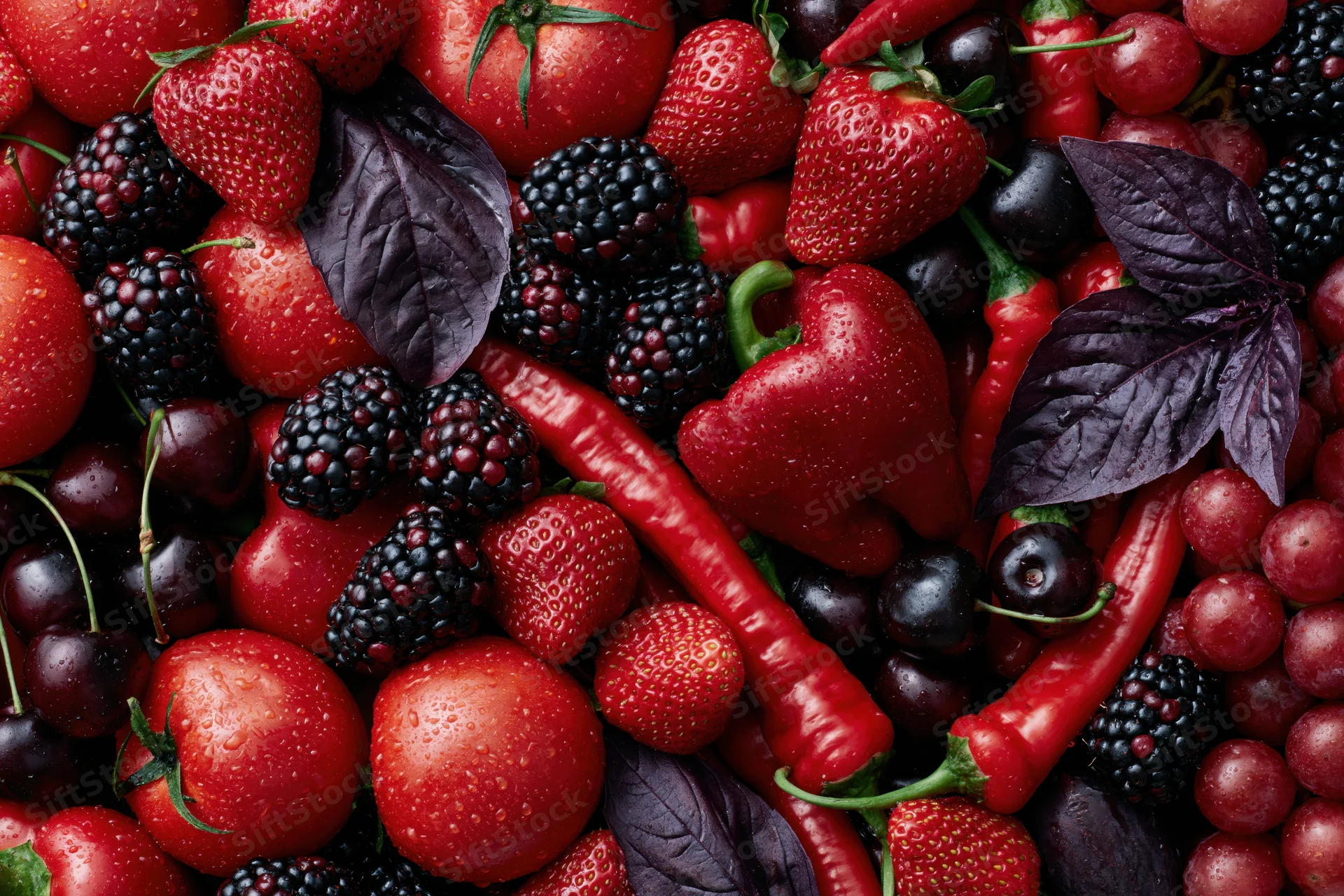 Overhead flat lay of mixed red and deep purple fruits and vegetables with basil leaves and water droplets