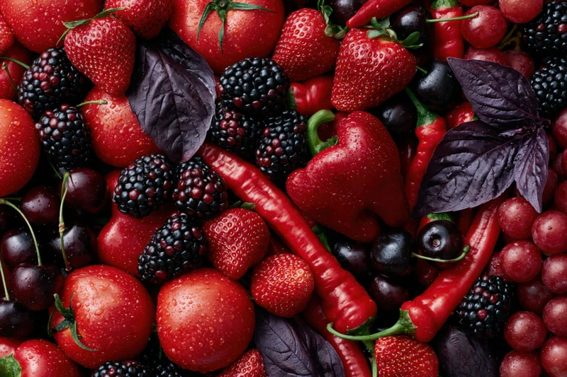 Overhead flat lay of mixed red and deep purple fruits and vegetables with basil leaves and water droplets