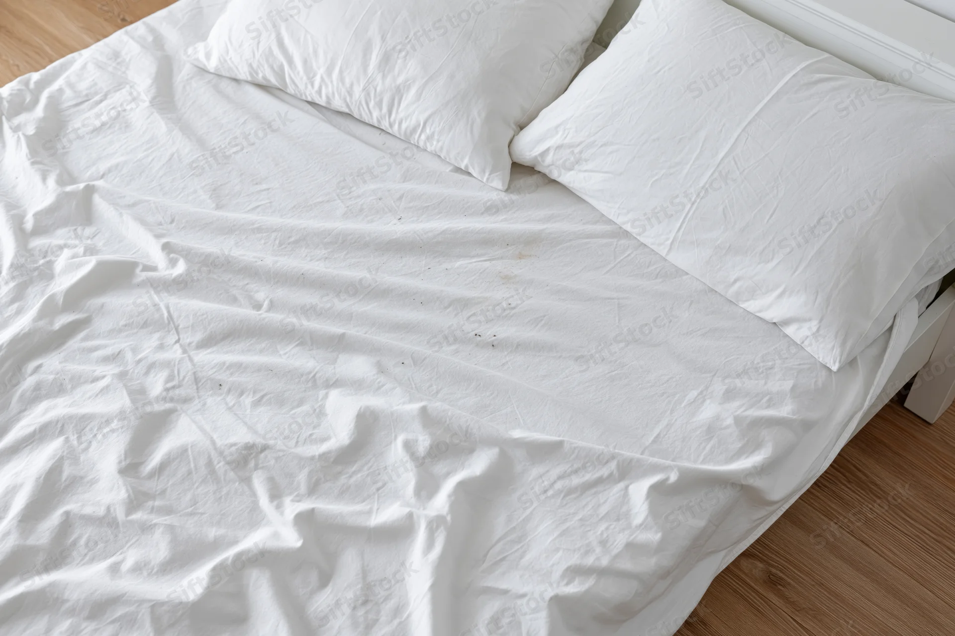 Unmade bed with crumpled white cotton sheets and pillows on wooden floor
