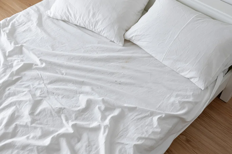Unmade bed with crumpled white cotton sheets and pillows on wooden floor