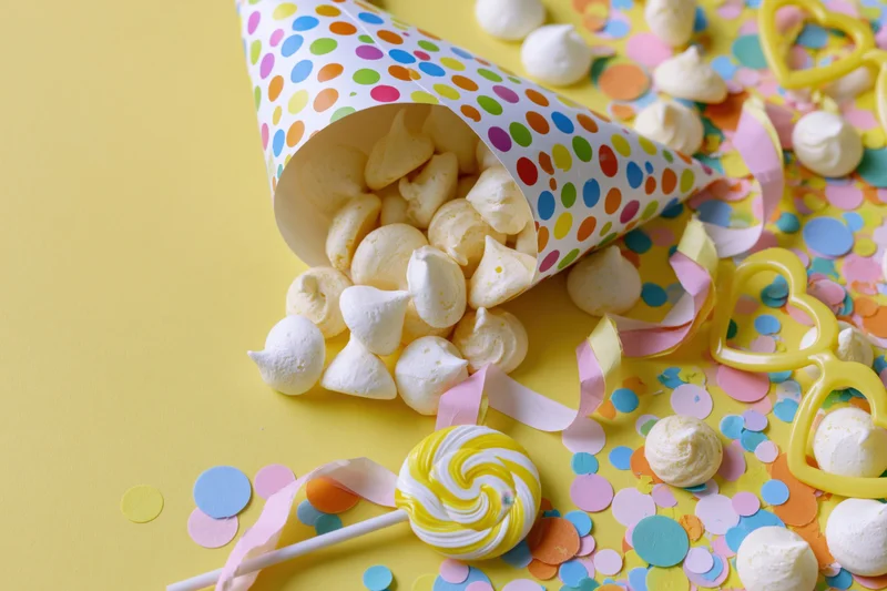 Party hat tipped over spilling marshmallows and confetti on yellow surface