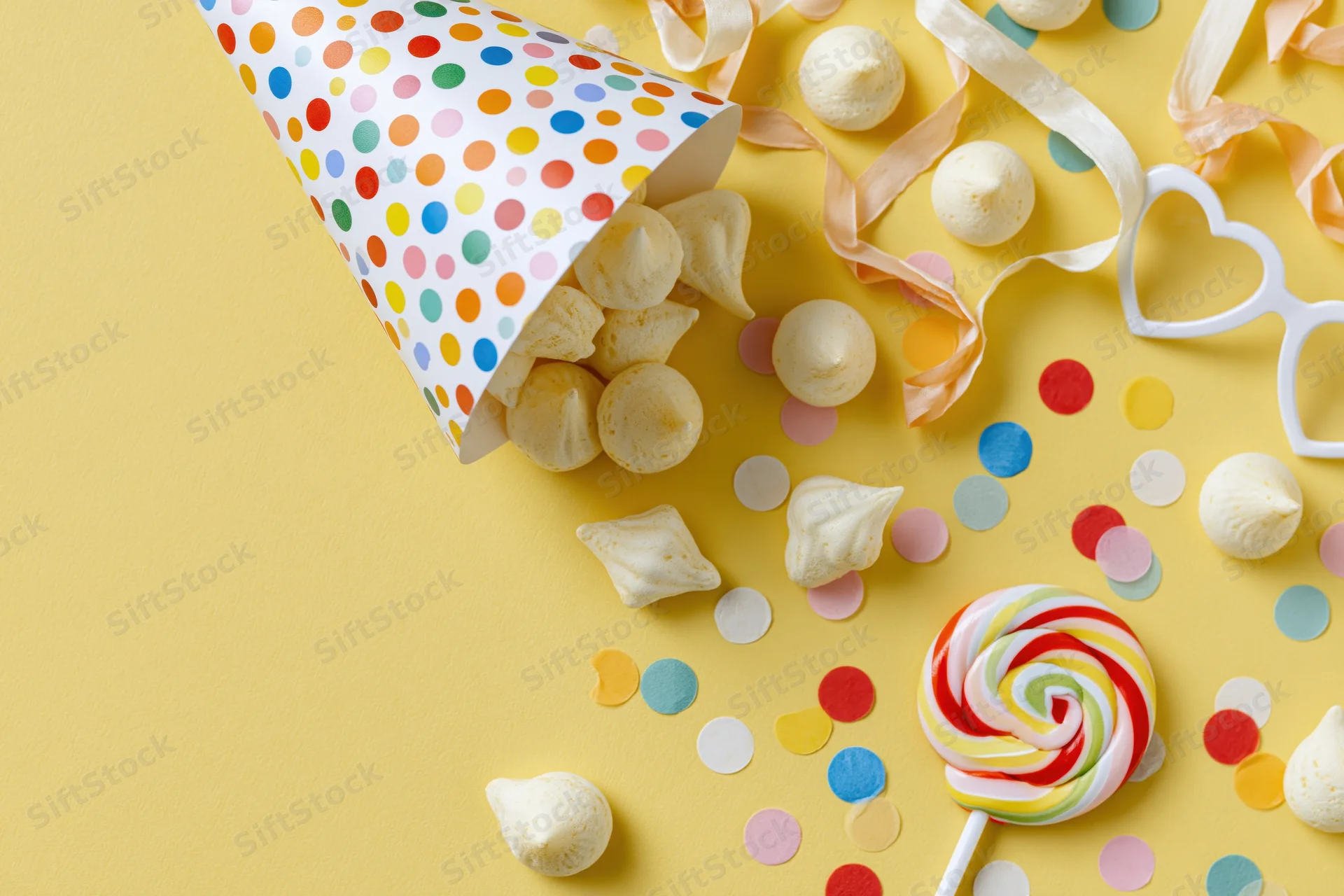 Party hat tipped over spilling meringues and confetti on yellow surface with lollipop and heart sunglasses nearby