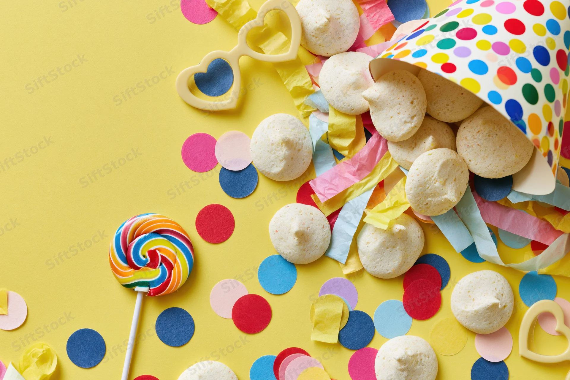 Party hat tipped over spilling confetti, candies, and streamers on yellow background