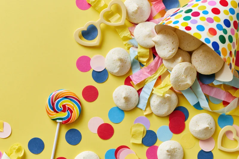 Party hat tipped over spilling confetti, candies, and streamers on yellow background