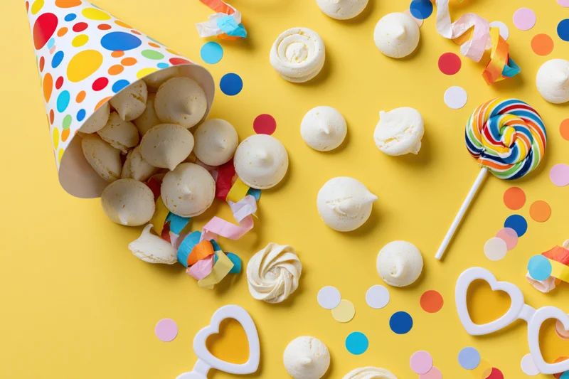 Party hat tipped over spilling meringues and candy on yellow surface with confetti and heart glasses