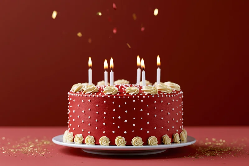 Red birthday cake with lit candles and confetti on a white stand against a red background