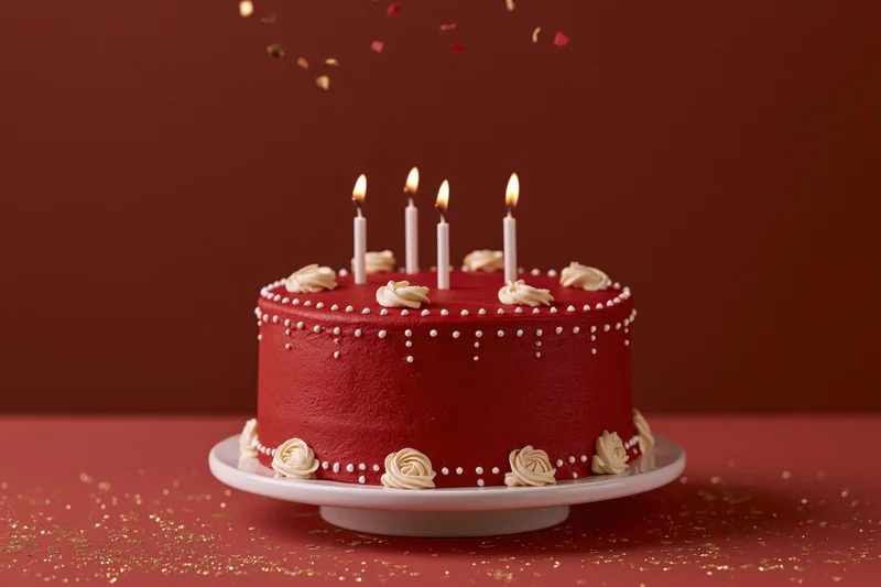 Red birthday cake with lit candles on white stand, confetti falling, gold glitter on surface