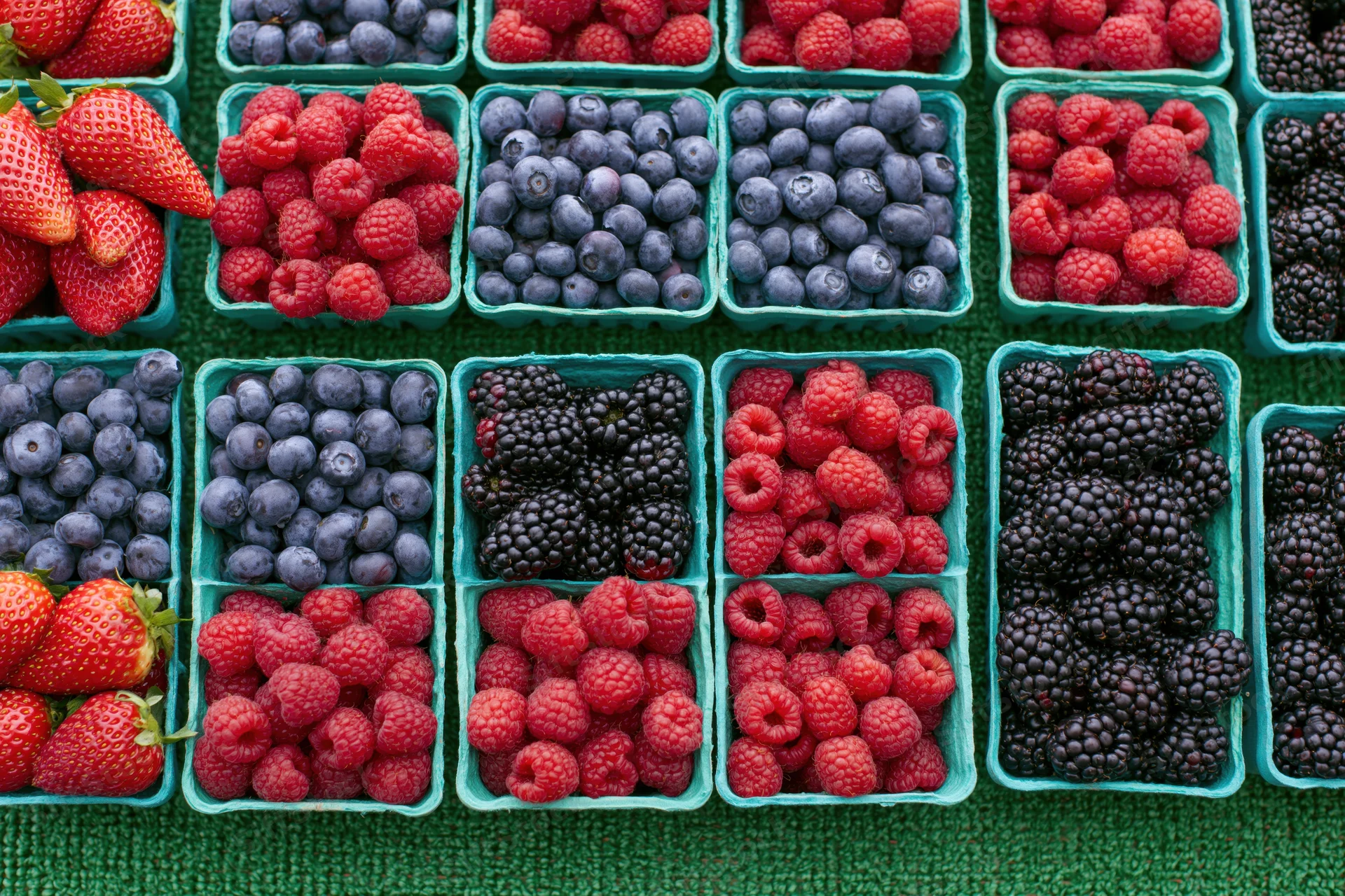 Rows of fresh strawberries blueberries raspberries and blackberries in green containers on a textured surface