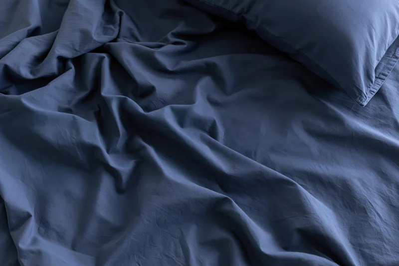 Rumpled dark navy blue sheets with a single pillow on an unmade bed