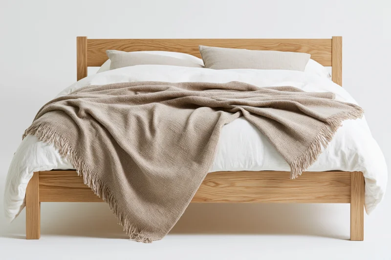 Double bed with light oak frame and white bedding, draped with beige blanket, in a minimalist studio setting