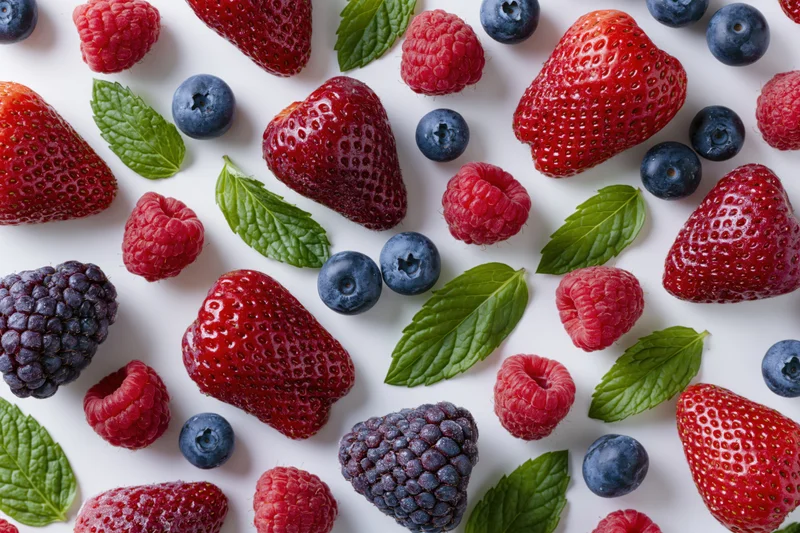 Scattered fresh strawberries raspberries blueberries and mint leaves on white surface