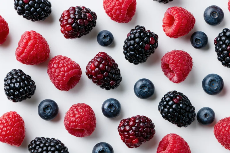 Scattered fresh raspberries blackberries and blueberries on white surface