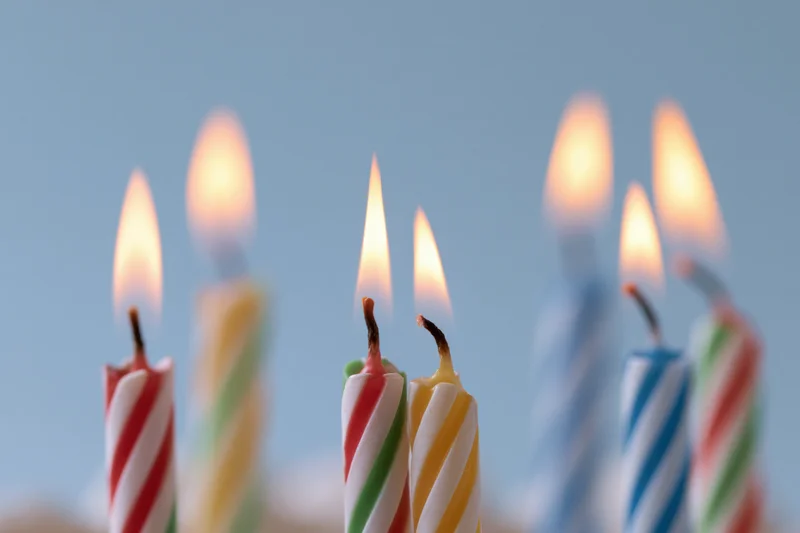 Lit birthday candles with spiral stripes in red, yellow, blue, and green arranged in a row against a soft blue background