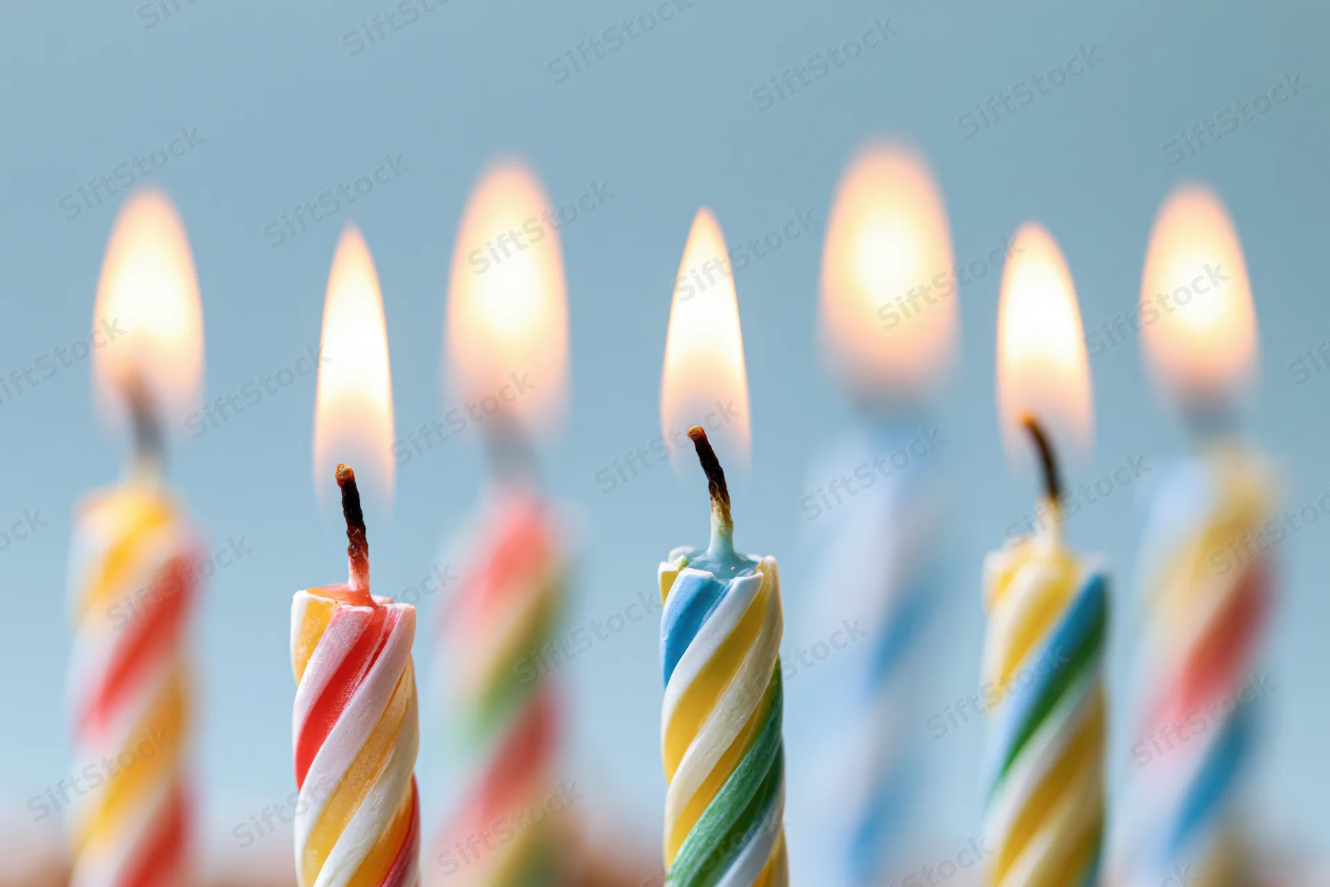 Lit birthday candles with spiral stripes in red yellow and blue, glowing against a soft blue background
