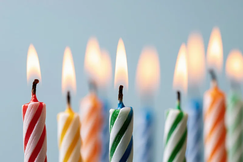 Lit birthday candles with spiral stripes in red, yellow, green, blue, and orange arranged in a row against a light blue background