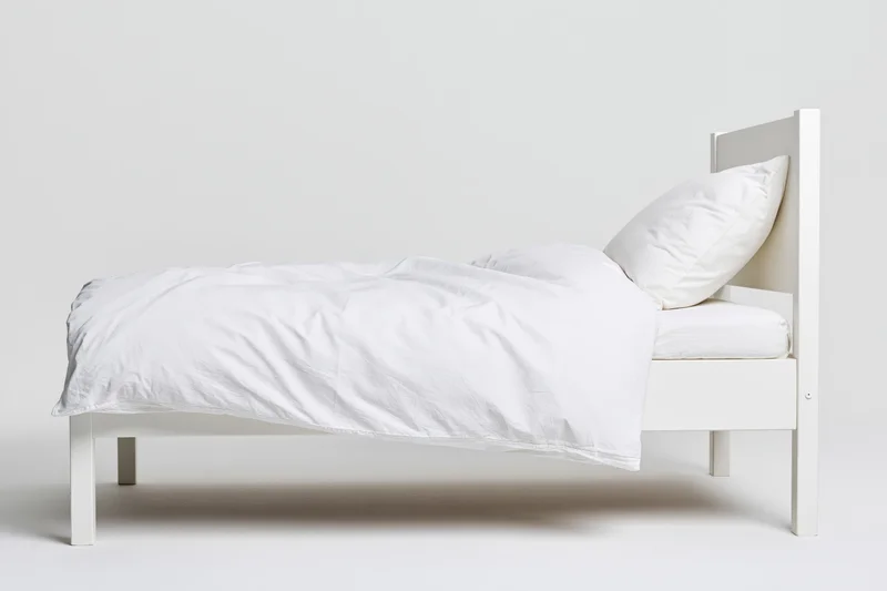 White wooden single bed with rumpled white bedding on plain background