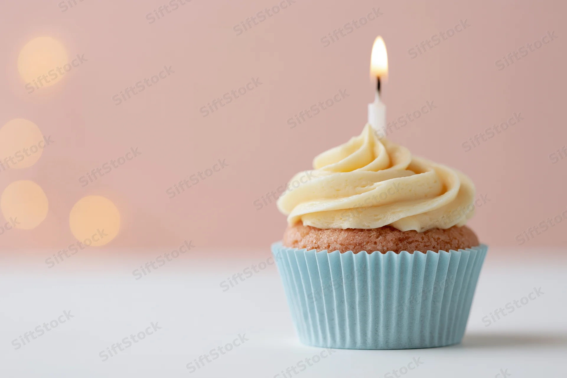 Single birthday cupcake with lit candle and vanilla frosting in pale blue paper liner on white surface with pink bokeh background