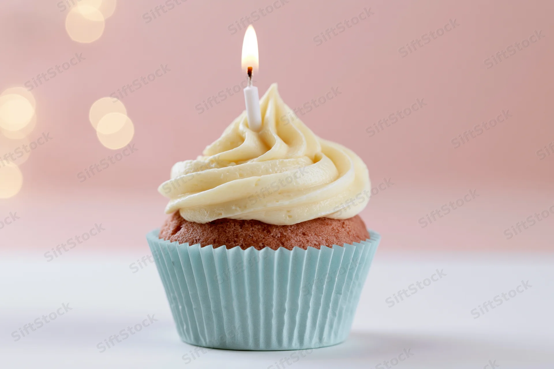 Single birthday cupcake with lit candle and vanilla frosting in pale blue paper liner on white surface with pink bokeh background