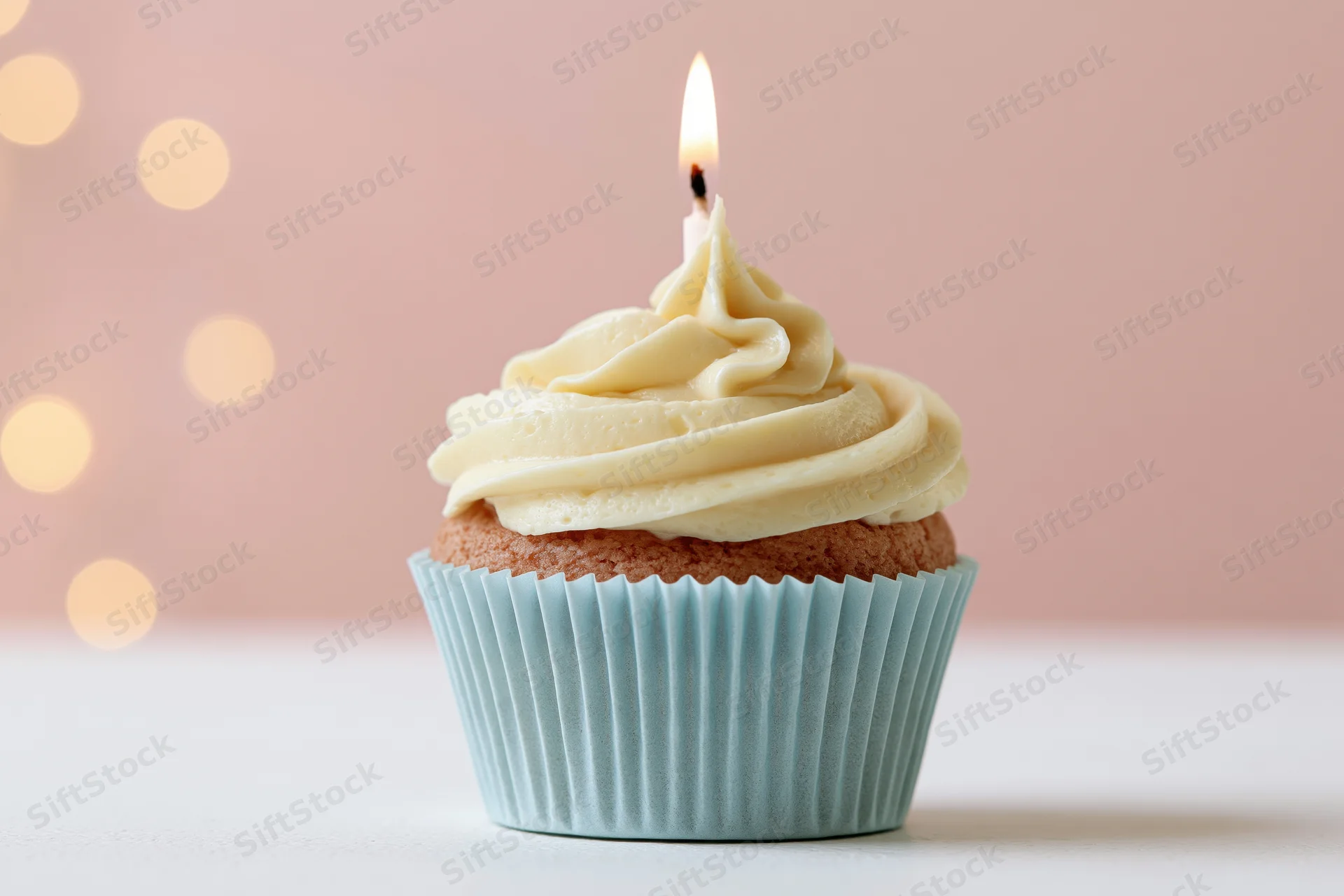Single birthday cupcake with lit candle and vanilla frosting in pale blue paper liner on white surface against pink bokeh background