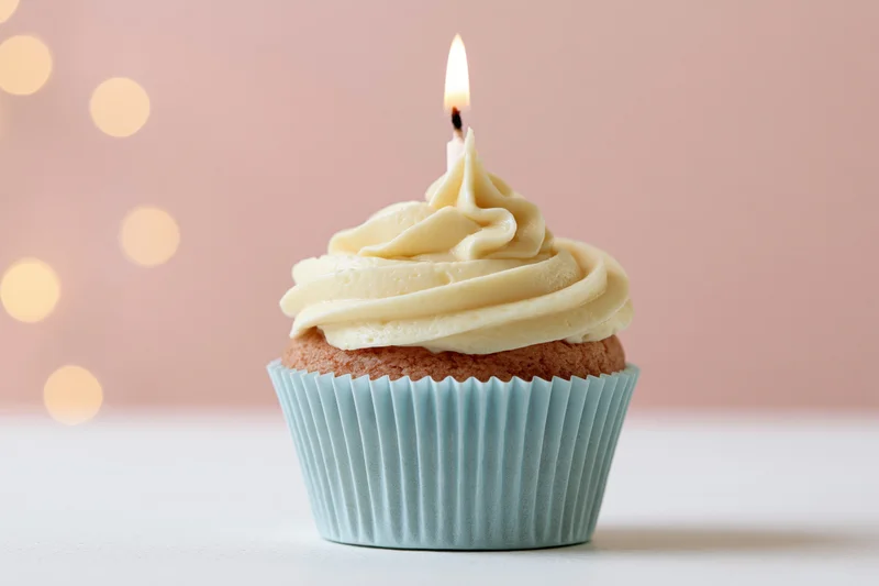 Single birthday cupcake with lit candle and vanilla frosting in pale blue paper liner on white surface against pink bokeh background