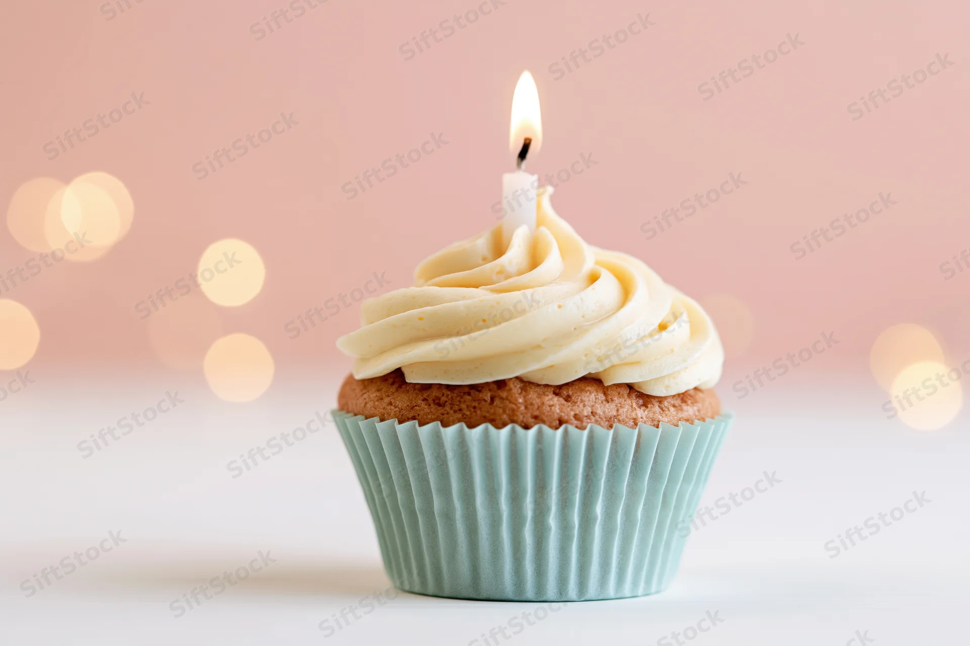 Single birthday cupcake with lit candle and vanilla frosting in pale blue paper liner on white surface with soft pink bokeh background