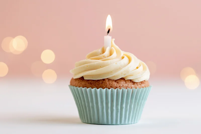 Single birthday cupcake with lit candle and vanilla frosting in pale blue paper liner on white surface with soft pink bokeh background
