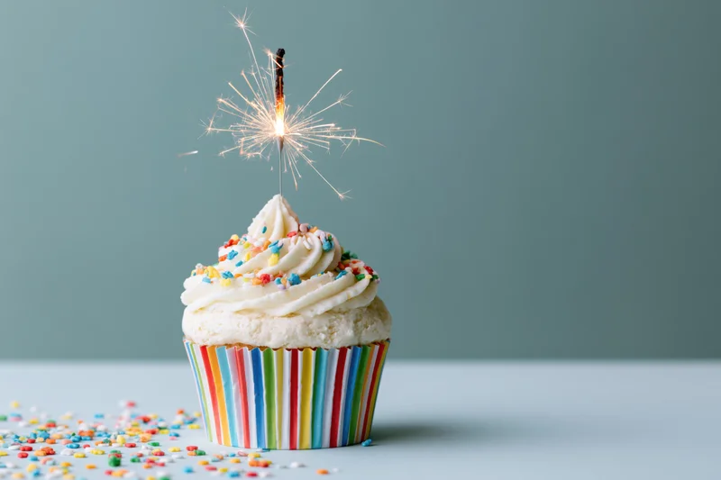 Single birthday cupcake with lit sparkler on top, surrounded by scattered sprinkles on a plain surface