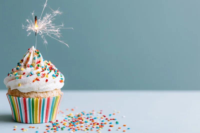 Single birthday cupcake with lit sparkler and colorful sprinkles on a blue background