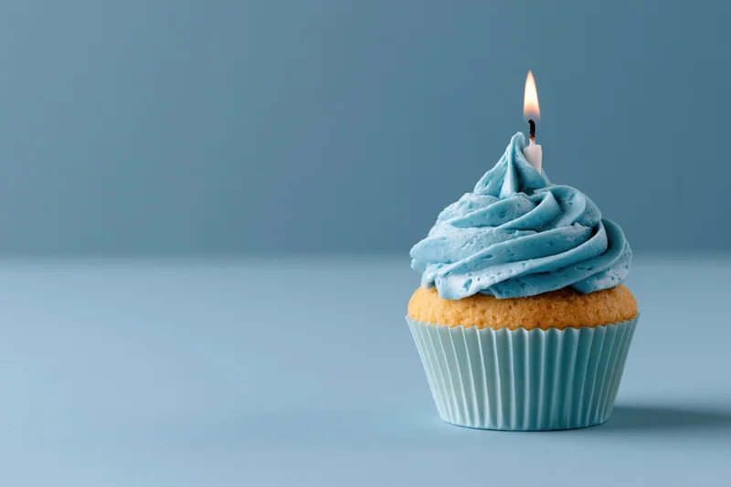 Single birthday cupcake with swirled blue frosting and lit candle on plain blue surface