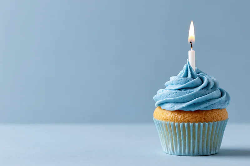 Single birthday cupcake with blue swirled frosting and lit candle on light blue surface