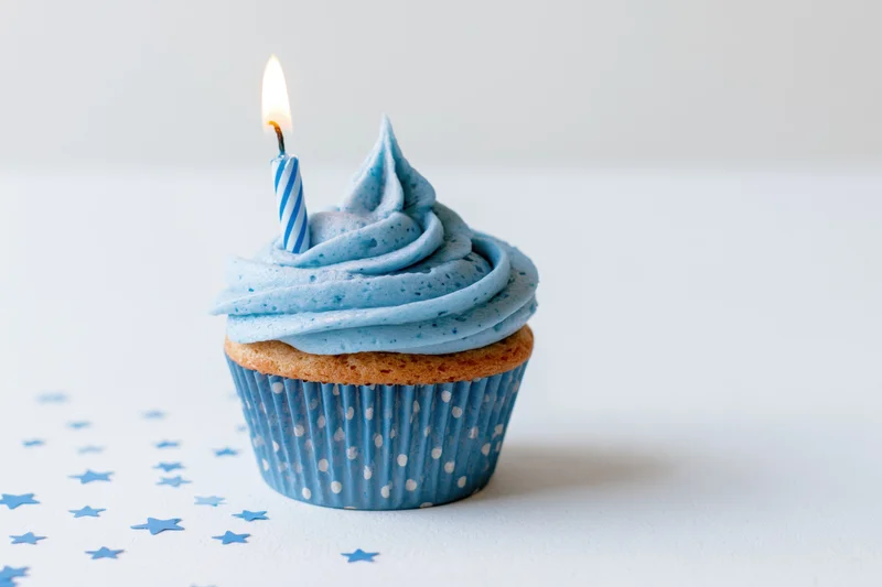 Single blue-frosted cupcake with lit candle on white surface surrounded by star confetti