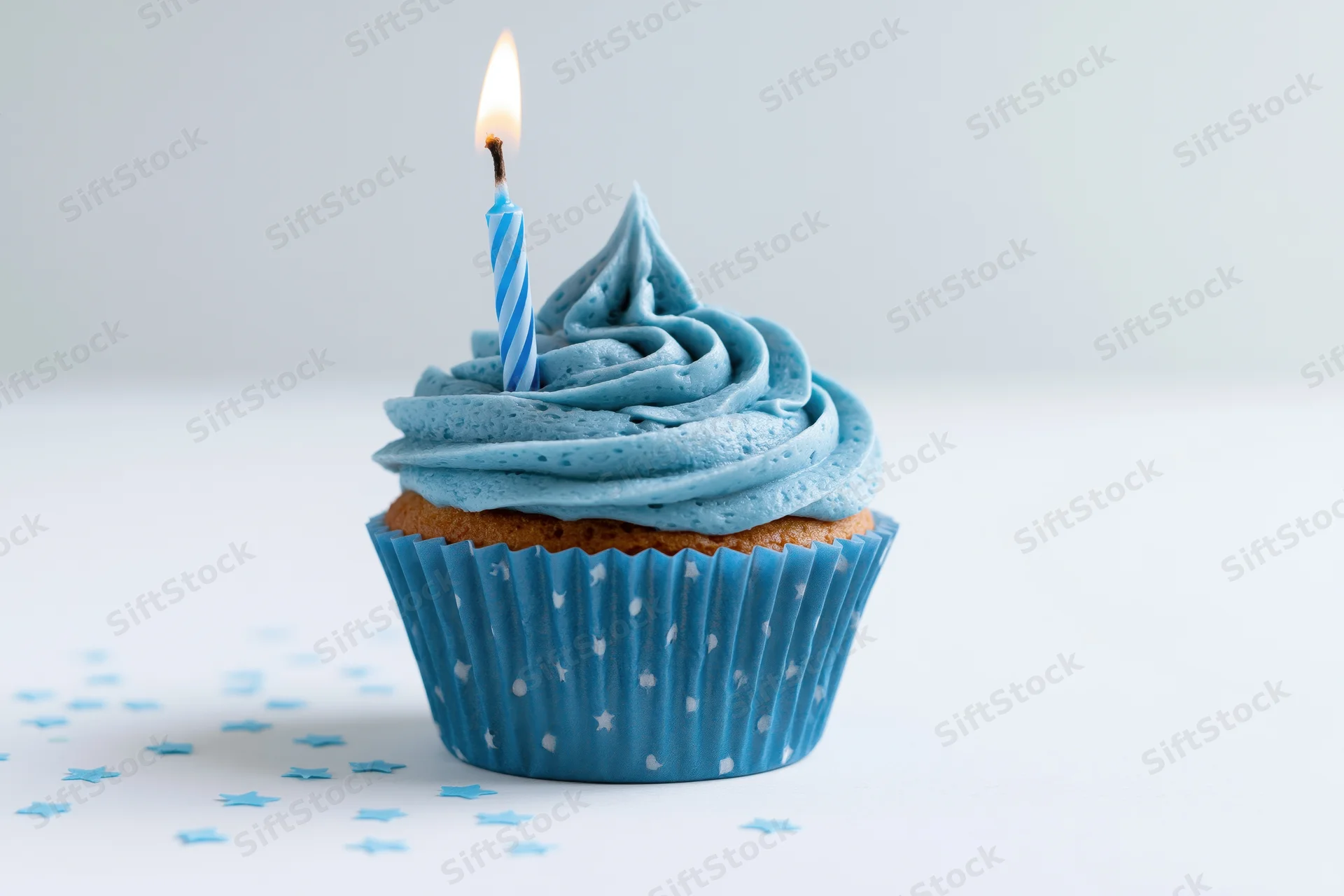 Single blue-frosted cupcake with lit candle on white surface surrounded by star confetti
