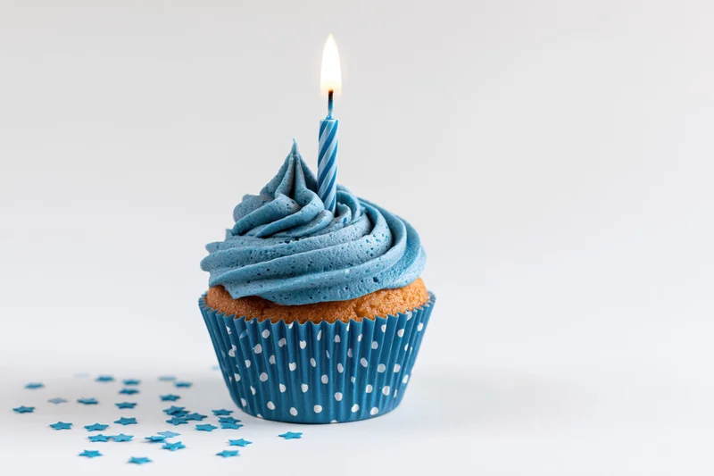 Single blue-frosted cupcake with lit candle and star confetti on white background