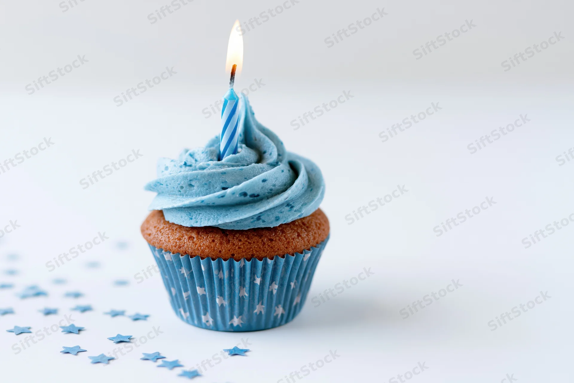 Single blue-frosted cupcake with lit candle on white surface surrounded by star confetti