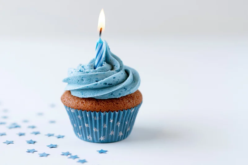 Single blue-frosted cupcake with lit candle on white surface surrounded by star confetti