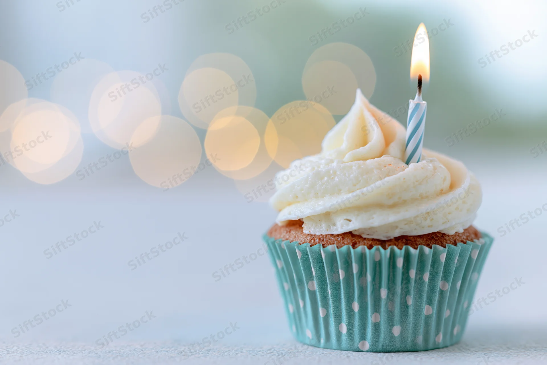 Single birthday cupcake with swirled white frosting and one lit candle in a blue polka dot paper liner on a light surface