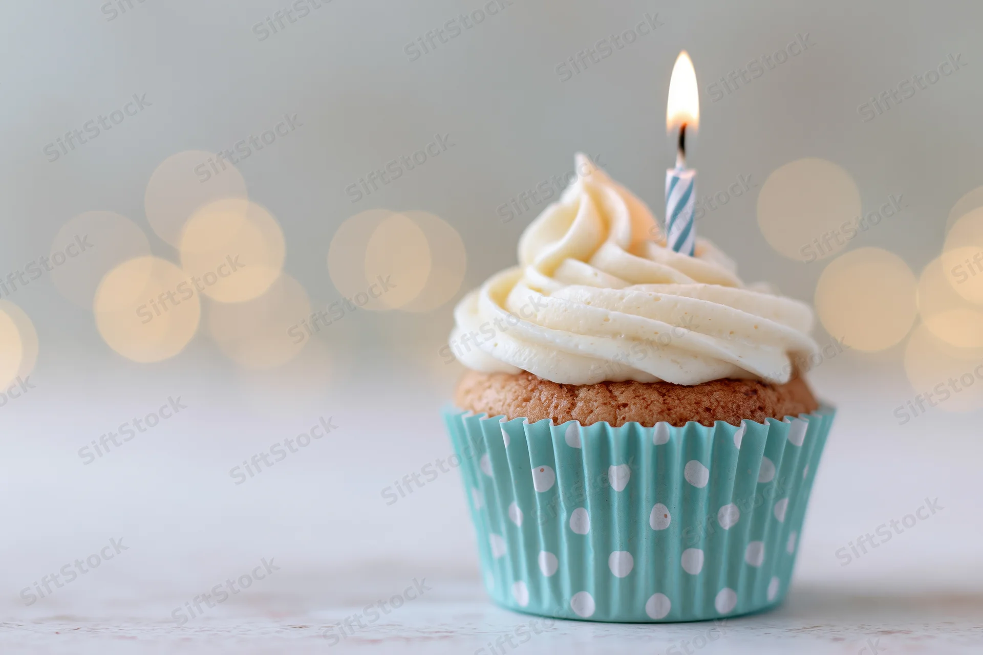 Single birthday cupcake with swirled white frosting and one lit candle in a blue polka dot liner on a light surface with blurred warm lights behind
