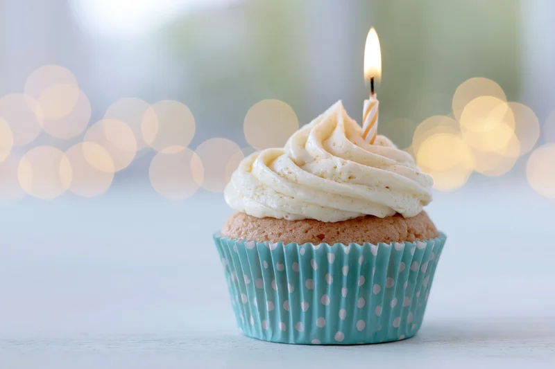 Single birthday cupcake with swirled white frosting and one lit candle in a blue polka dot paper liner on a light surface with bokeh background