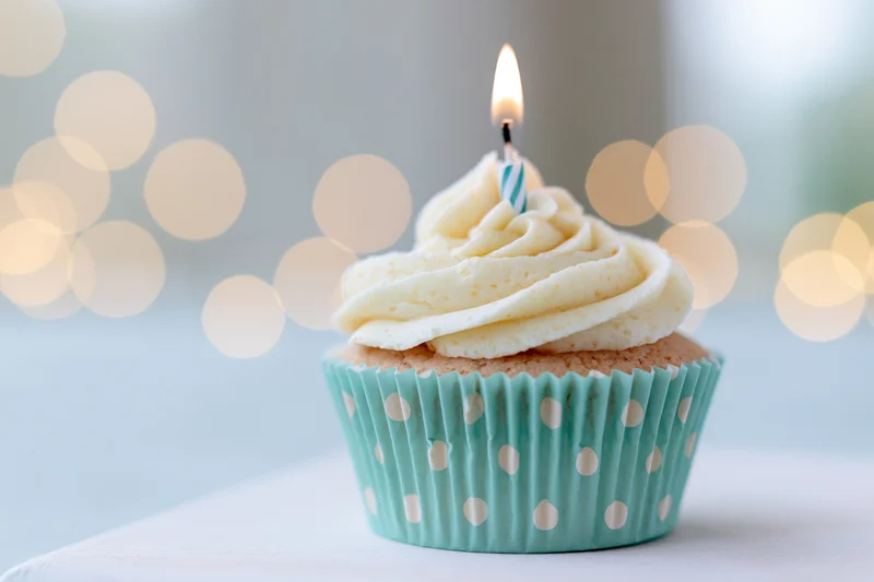 Single birthday cupcake with swirled white frosting and one lit candle in a blue polka dot paper liner on a white surface