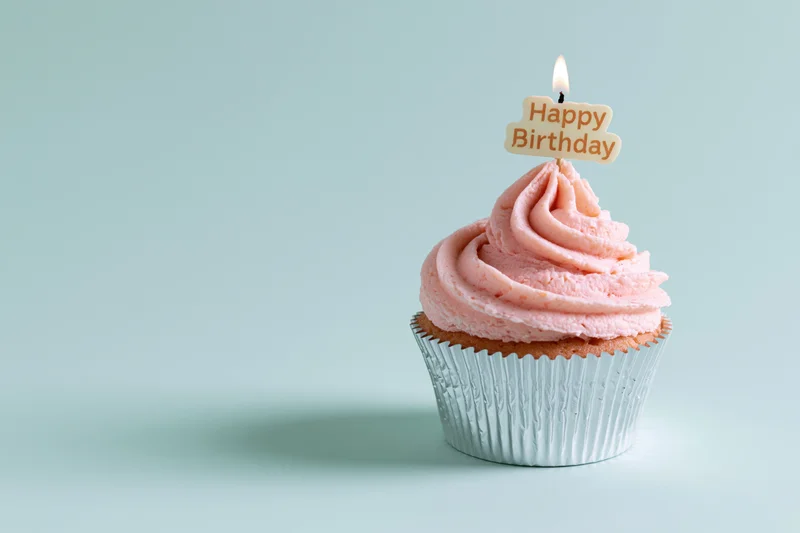 Single cupcake with pink frosting and lit birthday candle on a light blue surface