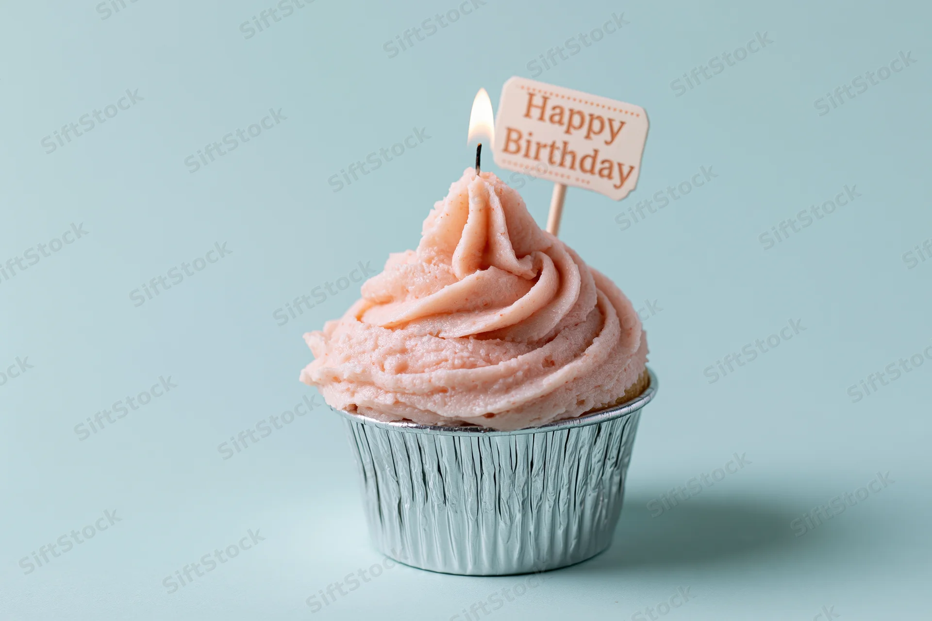 Single cupcake with pink frosting and lit candle in silver foil cup on light blue background