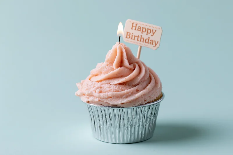 Single cupcake with pink frosting and lit candle in silver foil cup on light blue background