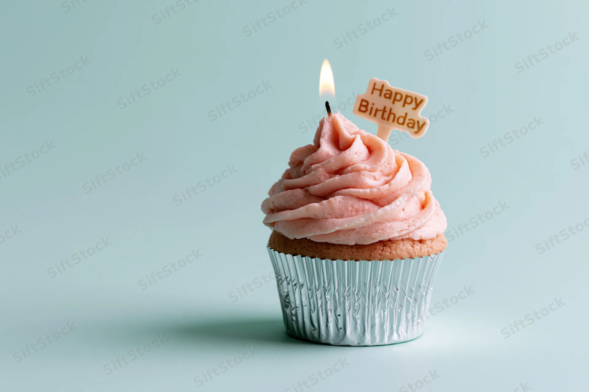 Single cupcake with pink swirled frosting and lit candle in silver foil wrapper on light blue background