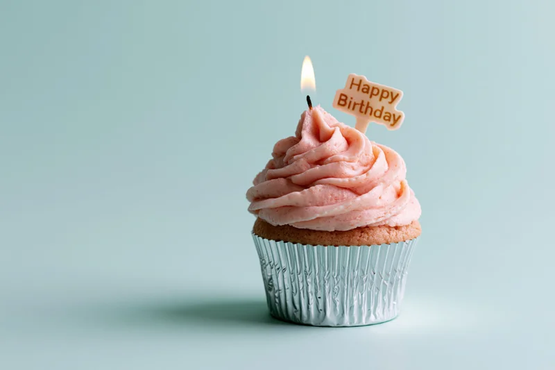 Single cupcake with pink swirled frosting and lit candle in silver foil wrapper on light blue background