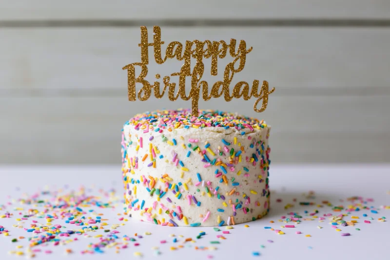 Small white frosted birthday cake with gold glitter topper and colorful sprinkles on white surface
