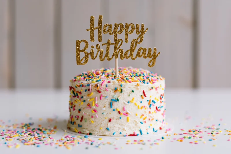 Small white frosted birthday cake with gold glitter topper and colorful sprinkles on a white surface