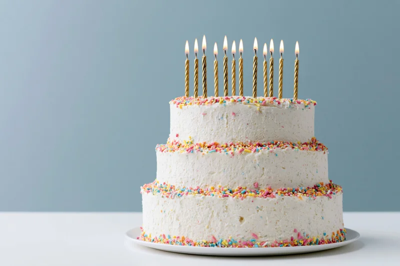 Three-tier white buttercream birthday cake with lit gold candles and colorful sprinkles on a white plate against a light blue background