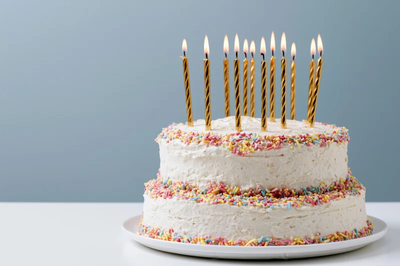 Two-tier white buttercream cake with colorful sprinkles and lit gold candles on a plate against a light blue background